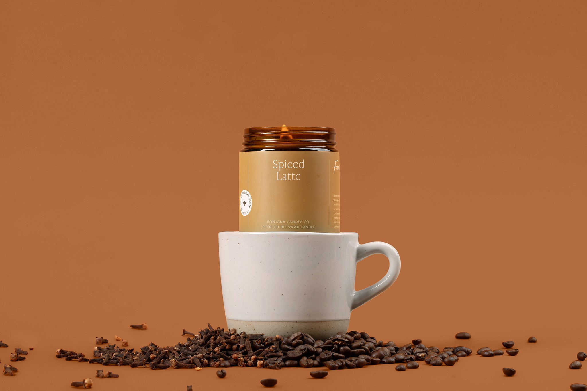 Lit amber Spiced Latte glass jar candle, sitting on top of a coffee mug, surrounded by coffee beans.