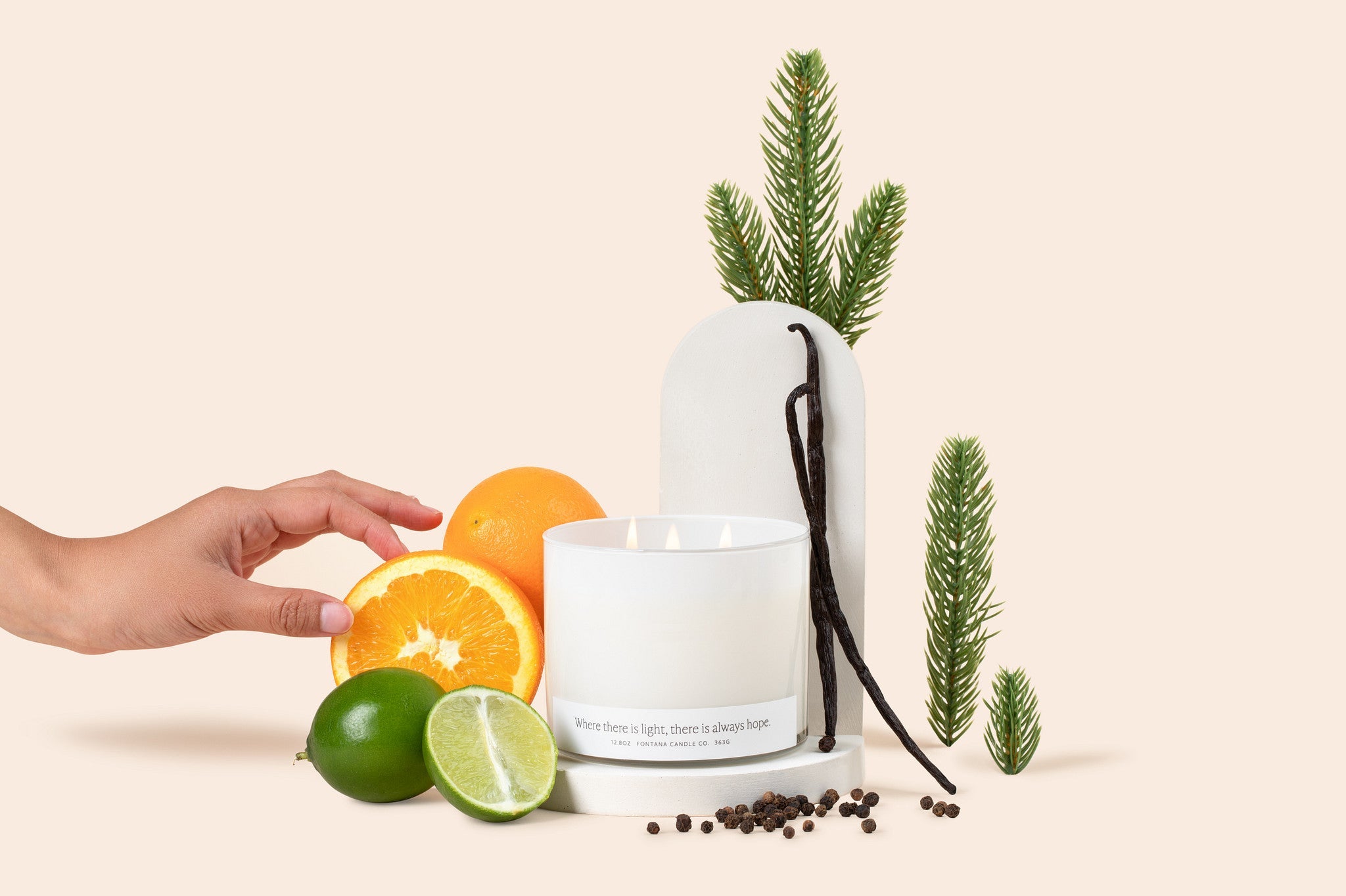 Lit 3-wick candle in a white glass vessel, surrounded by oranges, limes, and vanilla beans in front of a cream colored background.