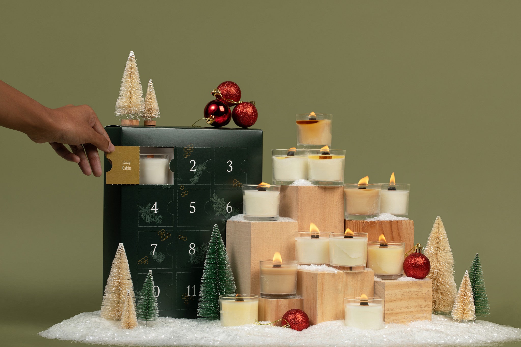 12 lit mini glass candles surrounded by snow, evergreen trees, and a green advent calendar box with the first window open, showing a mini candle in the first door.