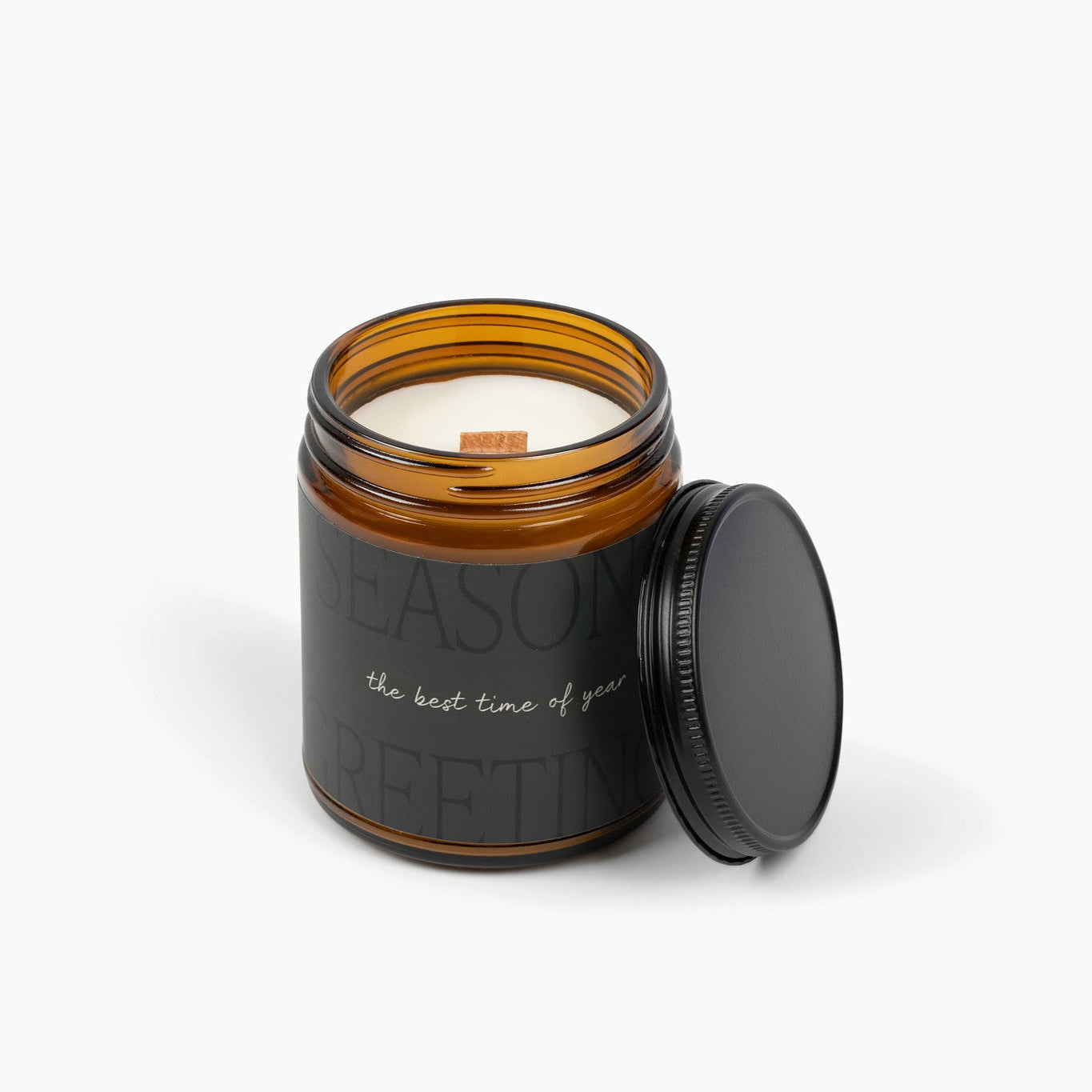 Unlit amber glass jar candle with an extremely dark green label that says: Season's Greetings: the best time of year and a wooden wick.