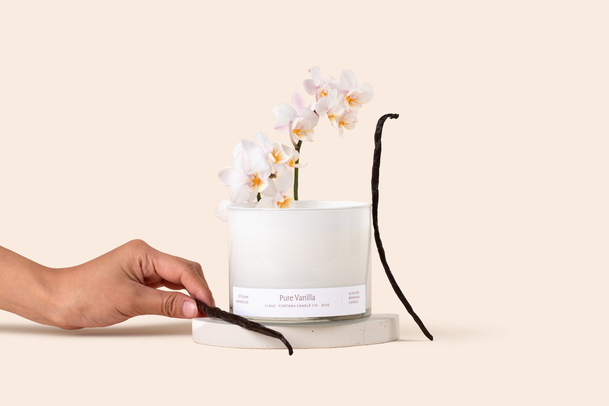 Vanilla 3 wick candle in a white glass vessel, surrounded by white orchids and vanilla beans, in front of a cream background.