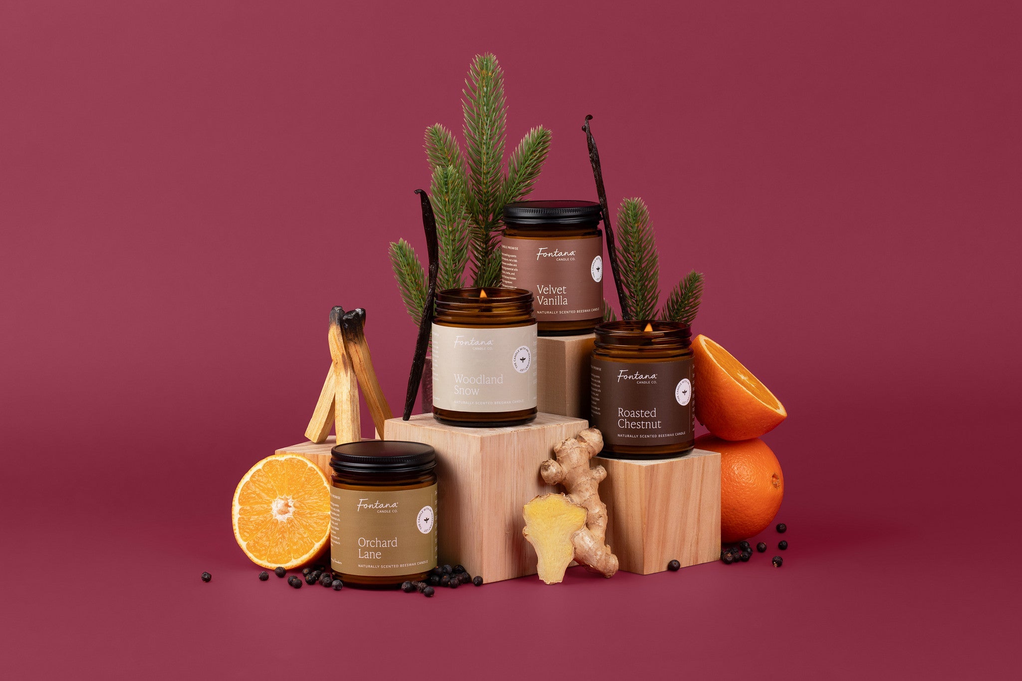 4 fall and holiday candles in amber glass jars, in front of a burgundy background, surrounded by spiced, oranges, and evergreen branches.