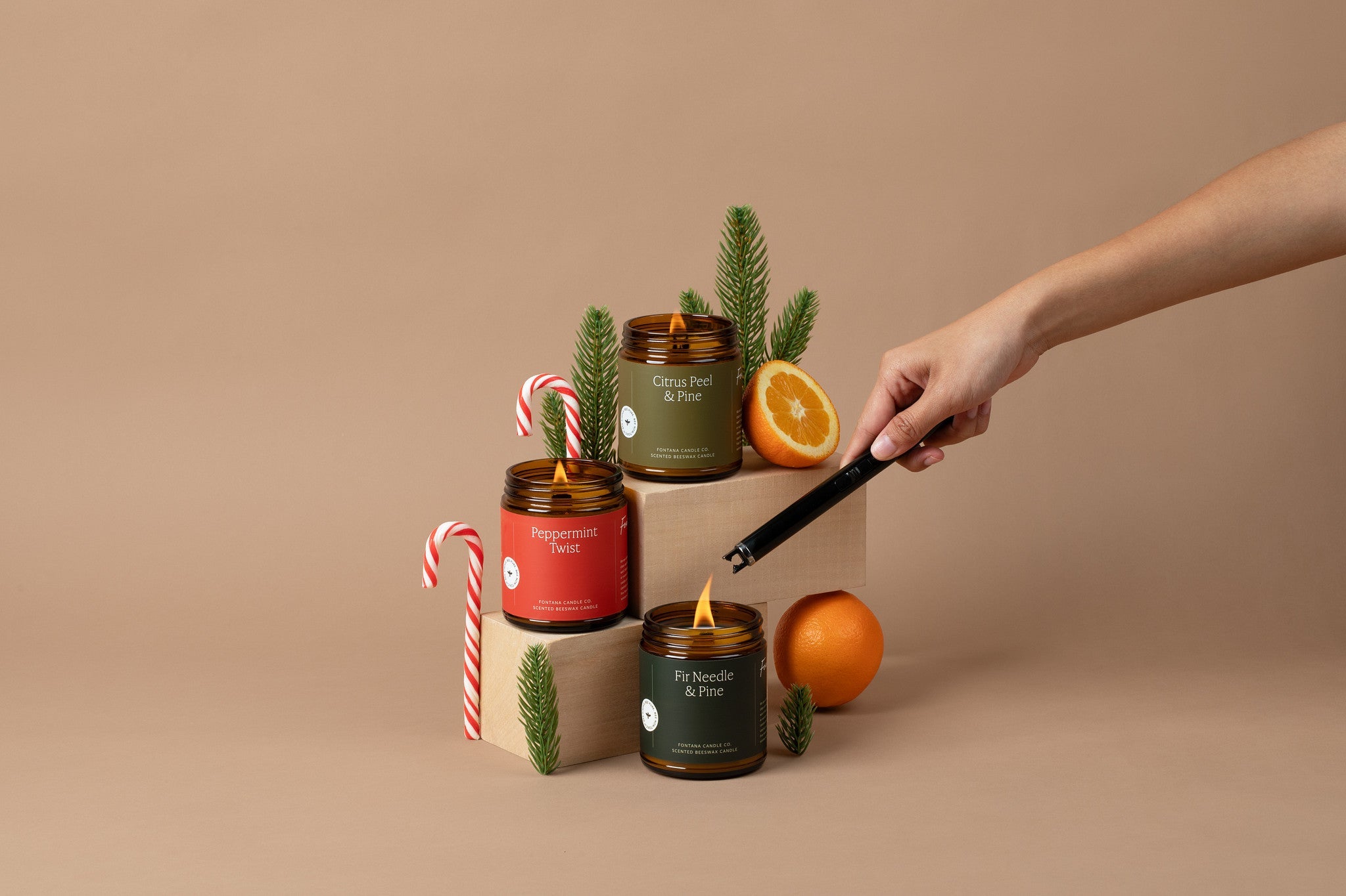 Three glass amber jar holiday candles on a beige background, surrounded by evergreen branches, candy canes, and oranges.