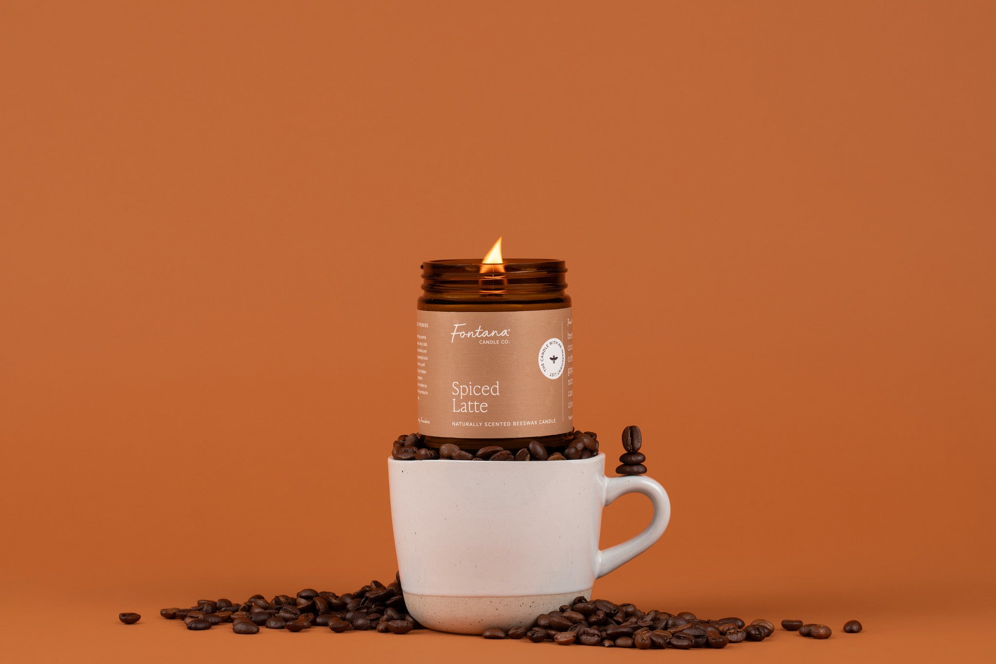 Lit Spiced Latte candle in an amber glass jar, sitting on top of a coffee mug that is full of coffee beans, in front of an orangeish brown background.