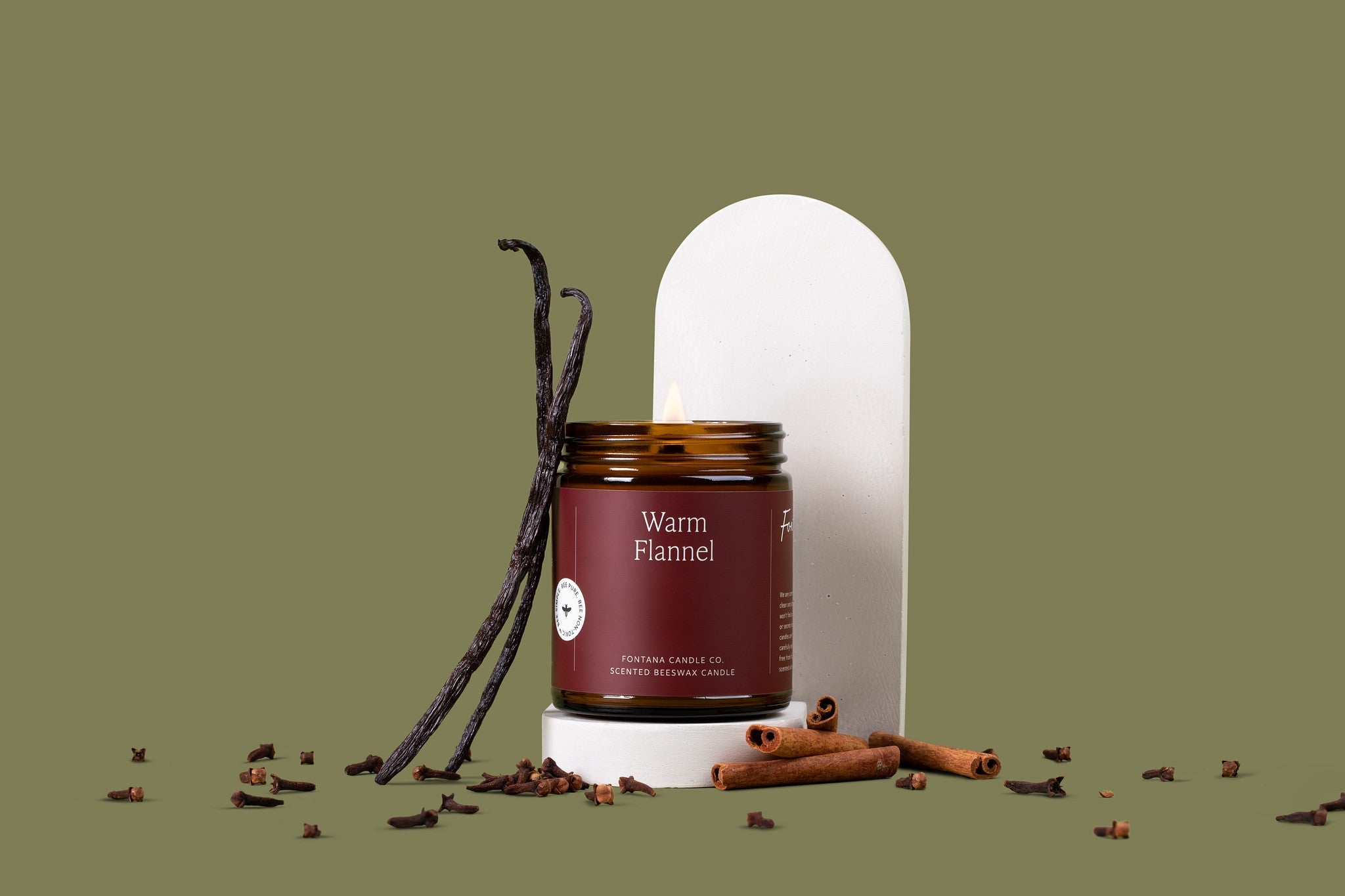 Lit amber glass jar candle with a deep maroon label, surrounded by vanilla beans, cloves, and cinnamon sticks, in front of a green background.