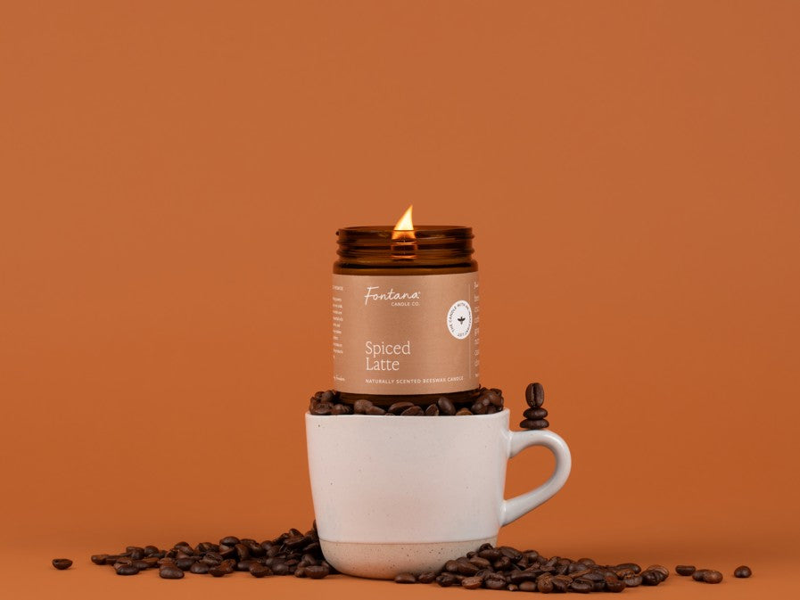 Lit amber glass jar candle, labeled Spiced Latte, sitting in a coffee mug, overflowing with coffee beans, in front of a orangish brown background.