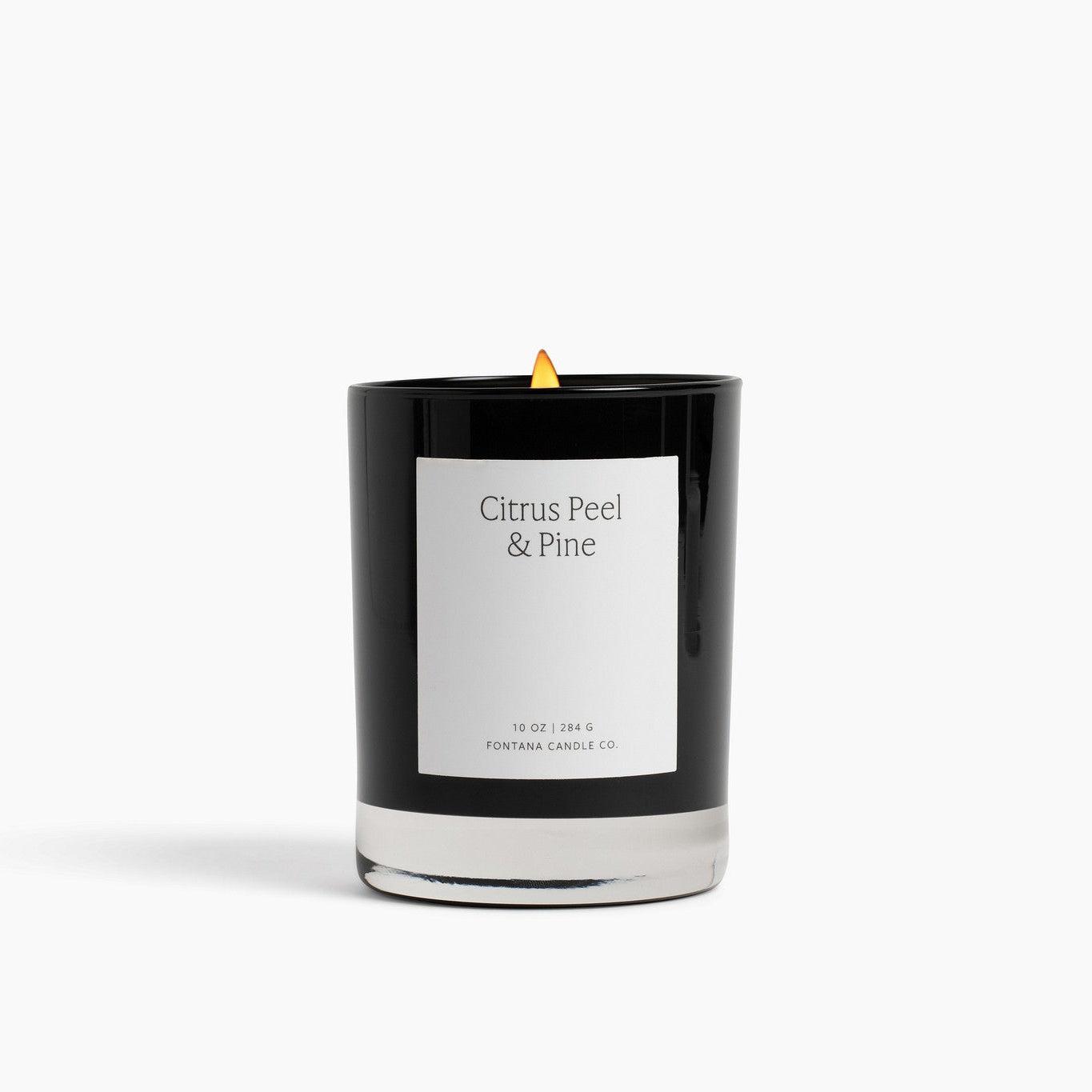 A black glass Noir candle with a white label reading 'Citrus Peel &amp; Pine' and a small yellow flame, photographed on a clean white background.