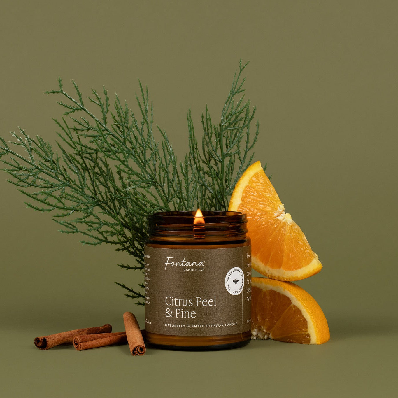 Candle with 'Citrus Peel &amp; Pine' scent surrounded by orange slices, cinnamon sticks, and greenery on a green background.