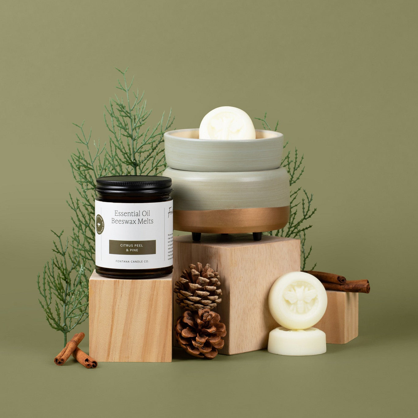 A styled product arrangement on a muted olive-green background featuring a jar labeled 'Essential Oil Beeswax Melt' alongside stacked ceramic wax melter, round wax melts, pinecones, cinnamon sticks, and evergreen branches.