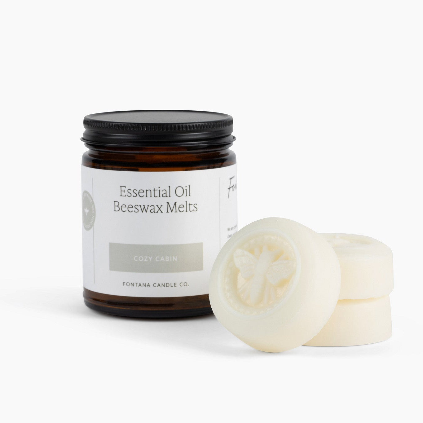 An amber glass jar labeled 'Essential Oil Beeswax Melts, Cozy Cabin' with a black lid, shown next to three round off-white beeswax melts on a clean white background.