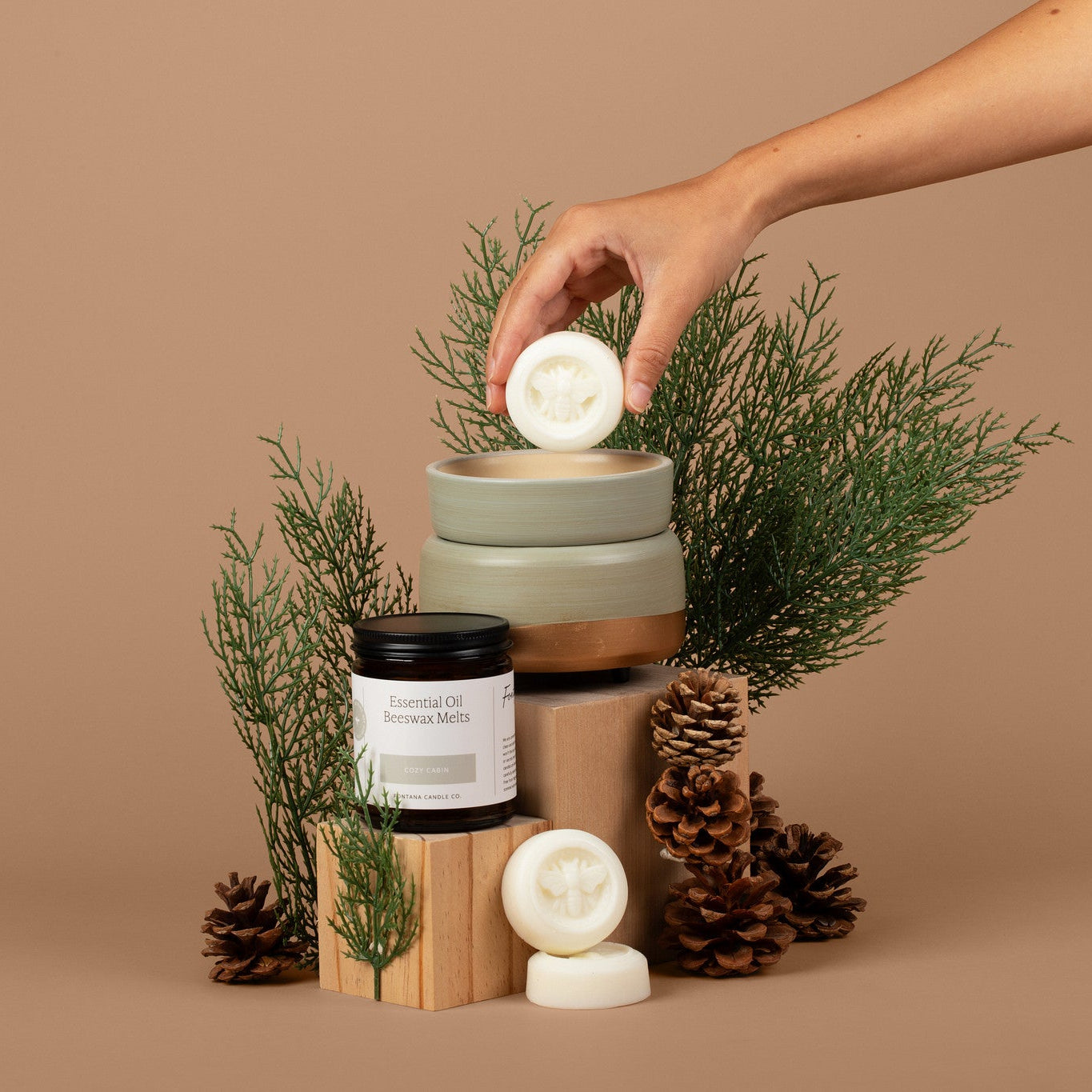 A hand places a round white wax melt into a wax melter on top of a stack of wooden blocks, styled with pine branches, pinecones, and a jar on a solid brown background.