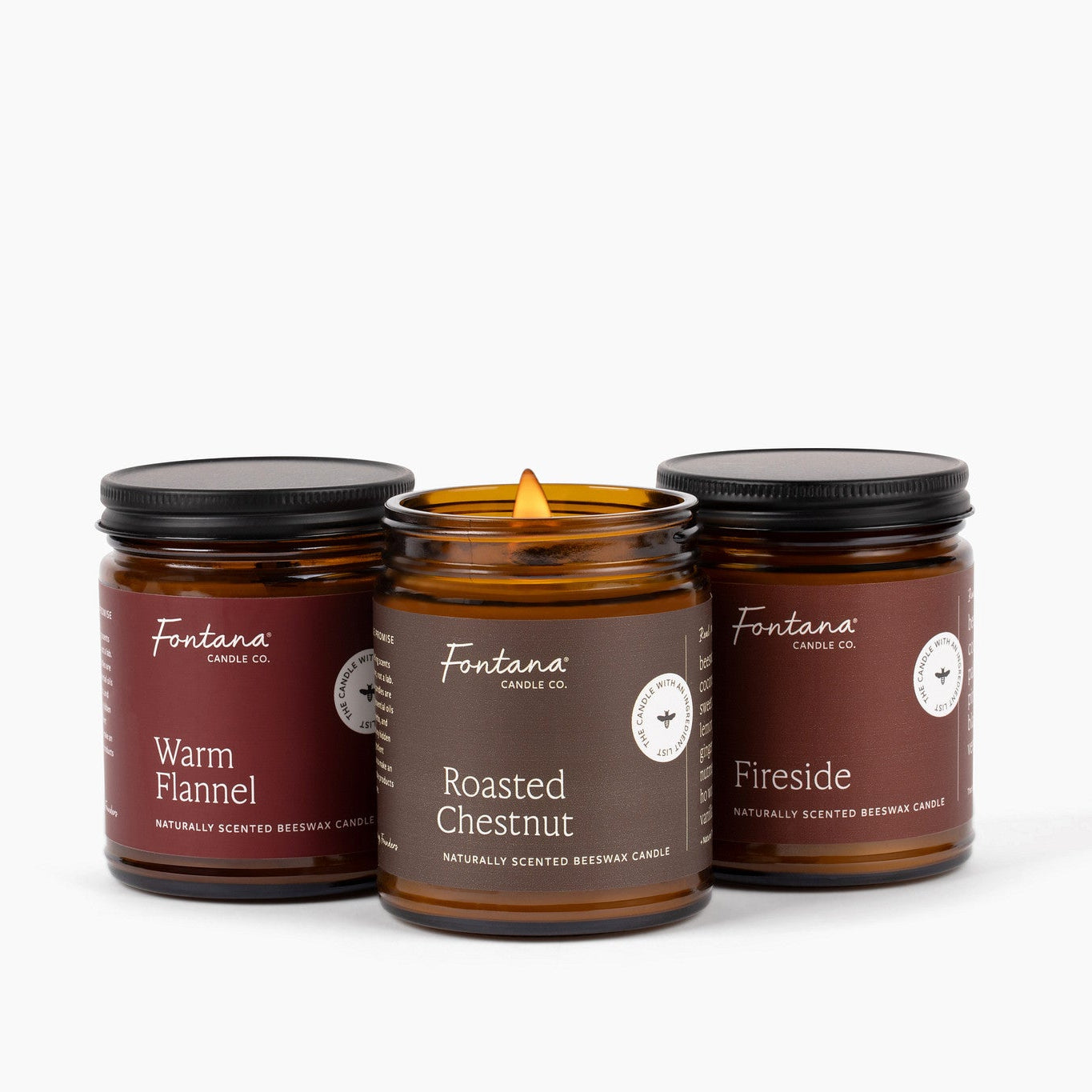 Three amber glass jar candles with black lids from Fontana Candle Co, Warm Flannel, Roasted Chestnut and Fireside are arranged on a white background. The center jar is open, showing a yellow flame, while the two side jars are closed.
