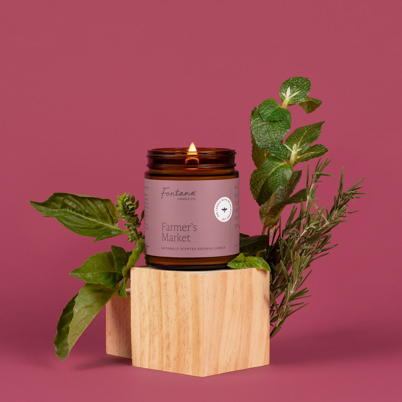 Amber glass jar candle labeled 'Farmer's Market' with greenery on a pink background.