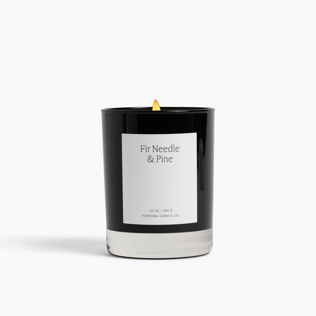 A black glass Noir candle with a white label reading 'Fir Needle &amp; Pine' and a small yellow flame, centered on a clean white background.