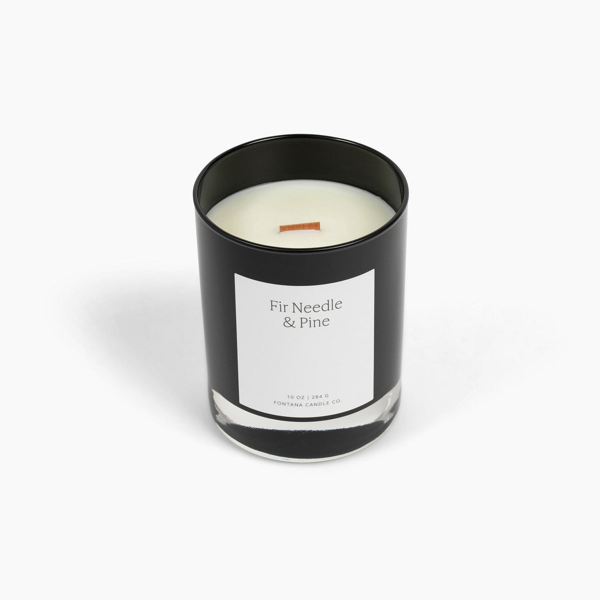 A black glass Noir candle with a white label reading 'Fir Needle &amp; Pine' and a wooden wick, centered on a clean white background.