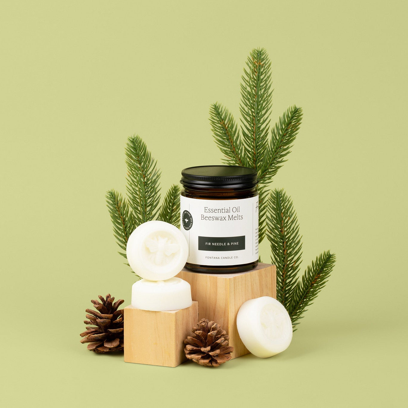 A jar of Essential Oils wax melts displayed on a wooden block with white wax melts, pinecones, and green pine branches against a light olive green background.