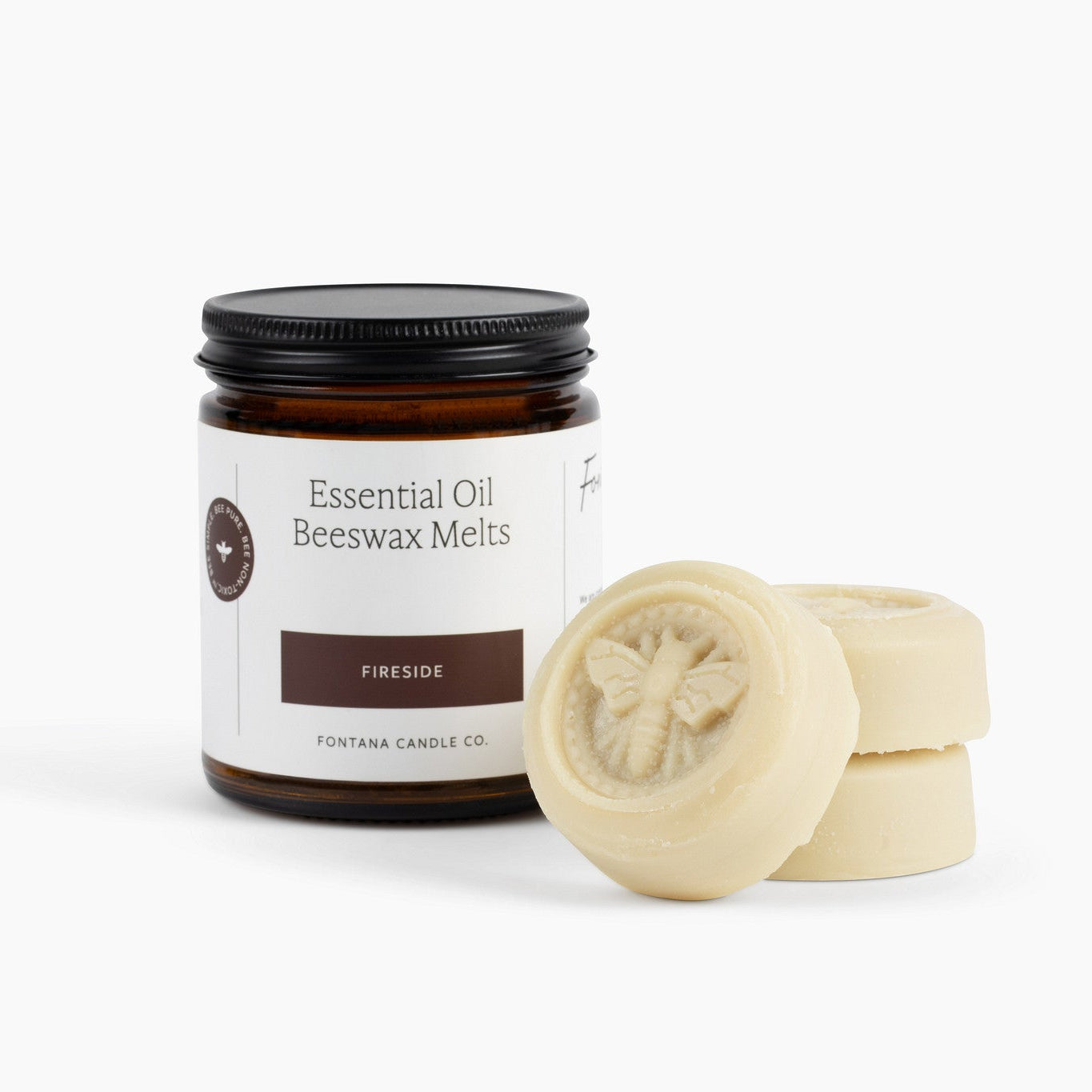 An amber glass jar labeled 'Essential Oil Beeswax Melts Fireside' with a black lid, shown next to three round tan beeswax melts on a clean white background.