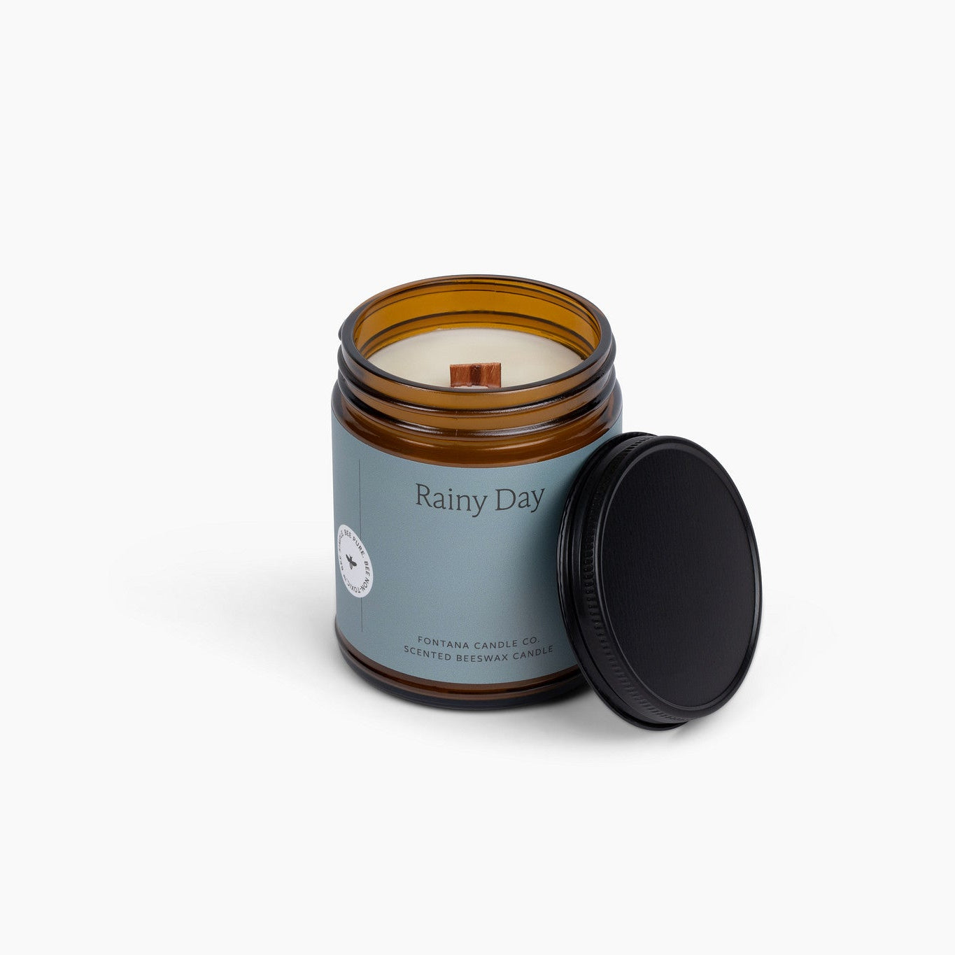 Amber glass jar candle with a blue label and wooden wick, in front of a white background.