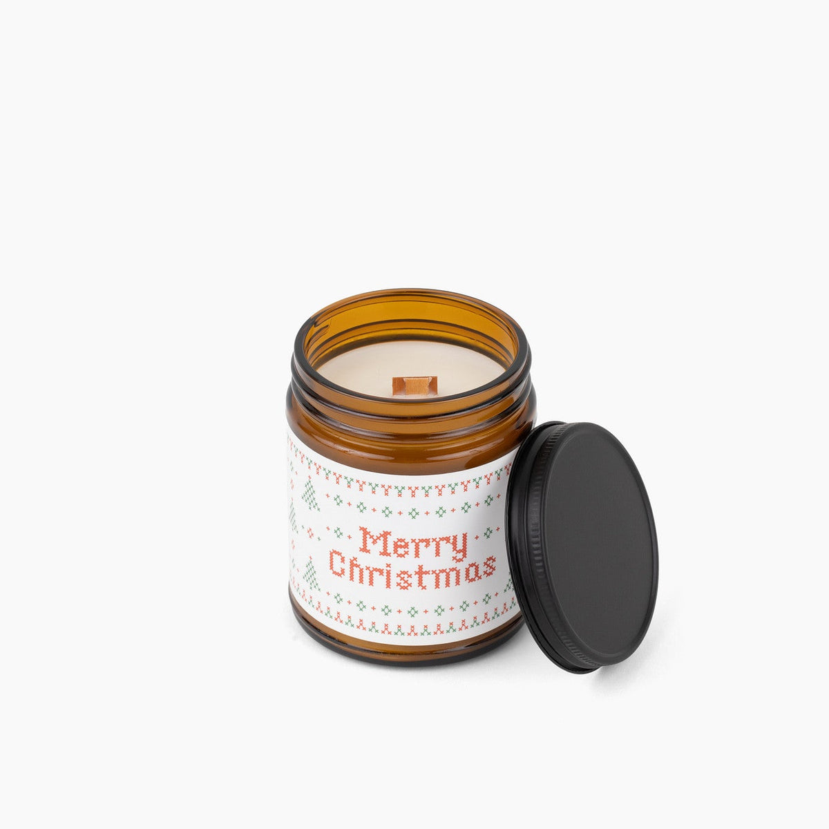 An open amber glass jar candle with a black lid resting beside it on a clean white background. The candle has a white label with red 'Merry Christmas' text and a light holiday pattern.