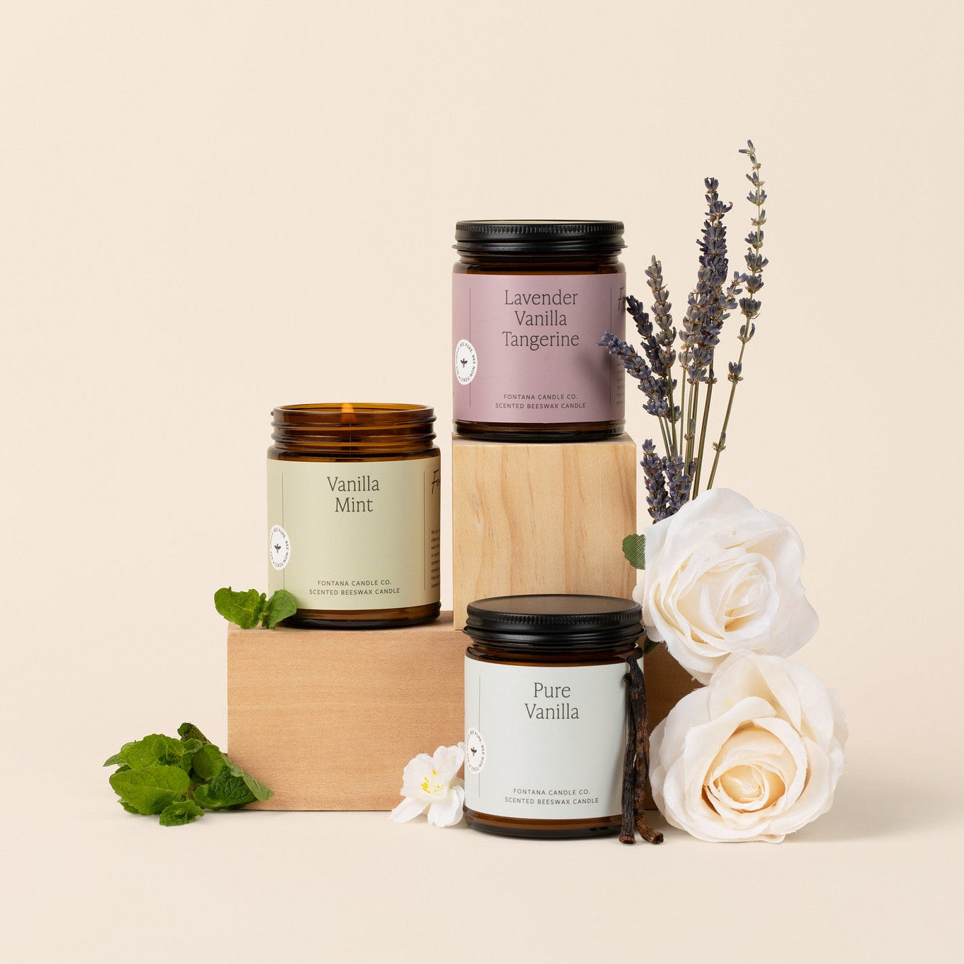 Three amber glass jar candles on a wooden block in front of a beige background surrounded by mint, vanilla, and flowers.