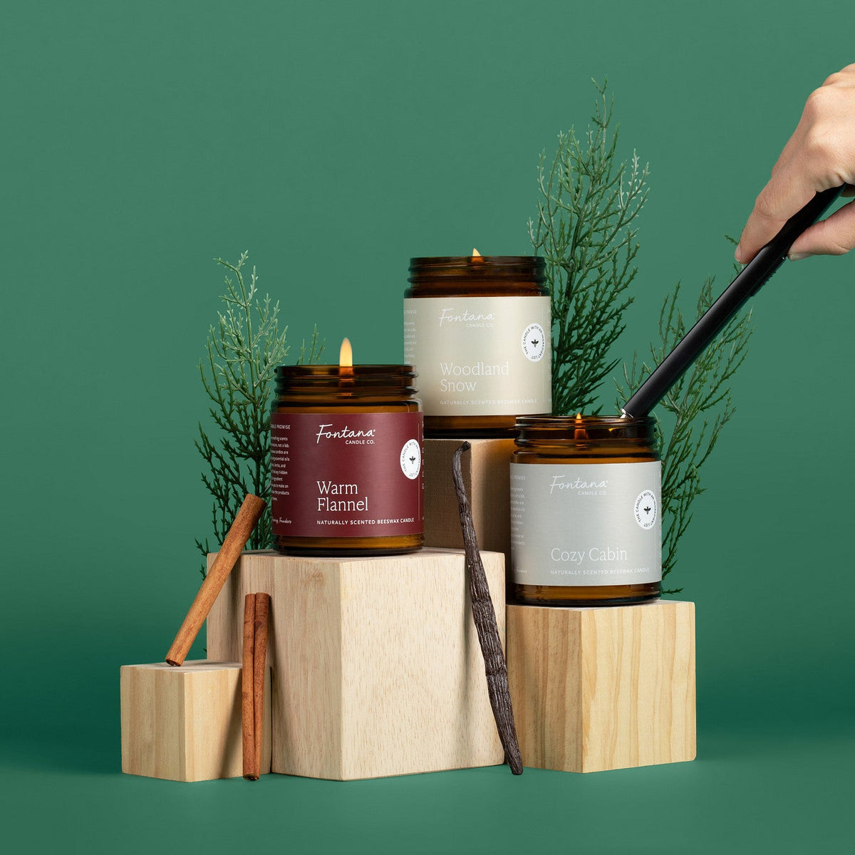 Collection of three amber glass jar candles on wooden blocks with greenery against a green background