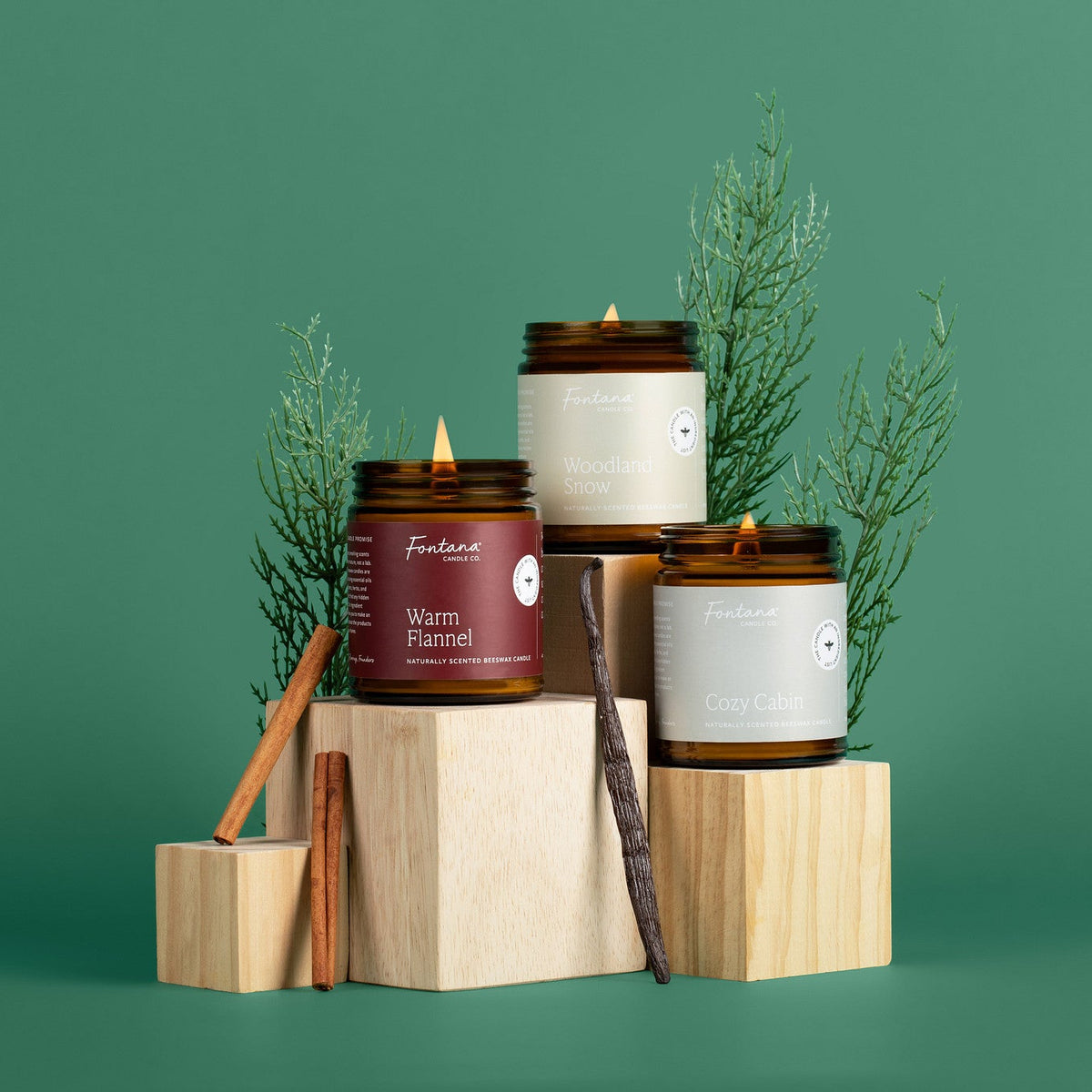 Three amber glass jar candles on wooden blocks with greenery against a green background