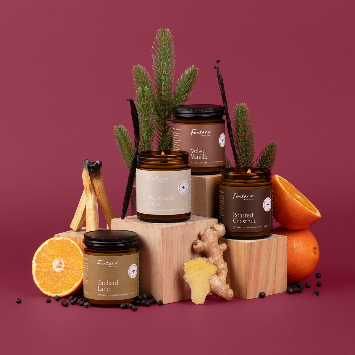 Four jar amber candles named Orchard Lane, Woodland Snow, Velvet Vanilla and Roasted Chestnut on a crimson background surrounded by fir needle, vanilla beans, oranges, ginger and juniper berries
