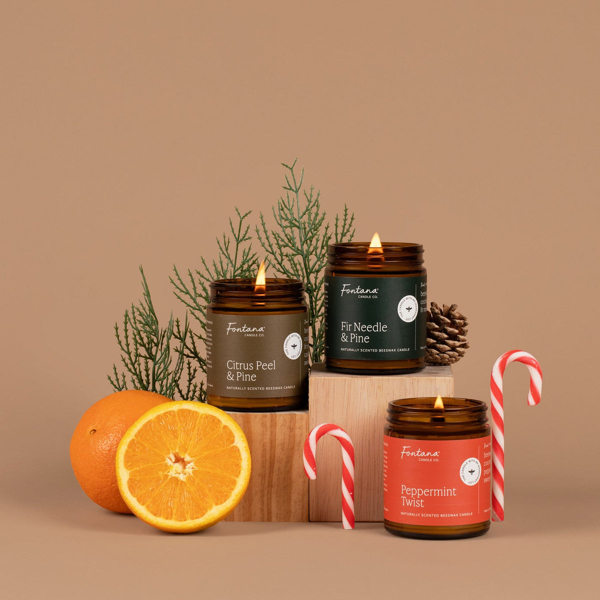 Three candles in glass jars with different scents on a brown background, surrounded by oranges and candy canes.