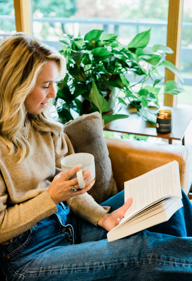 Nicole Jaques reading a book and holding a mug in a cozy living room, with a blurred Homewood candle burning in the background.