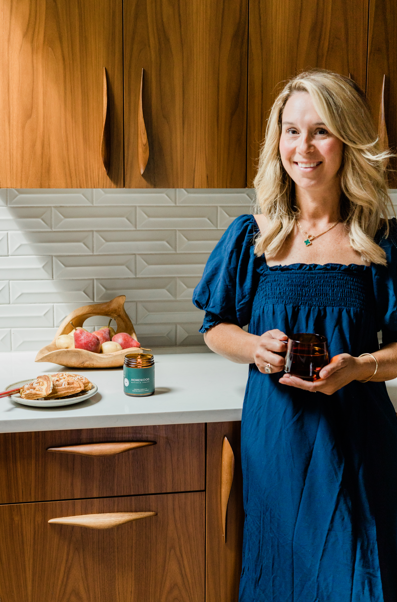 Nicole Jaques in a blue dress holding a mug in a kitchen, next to a burning Homewood candle.