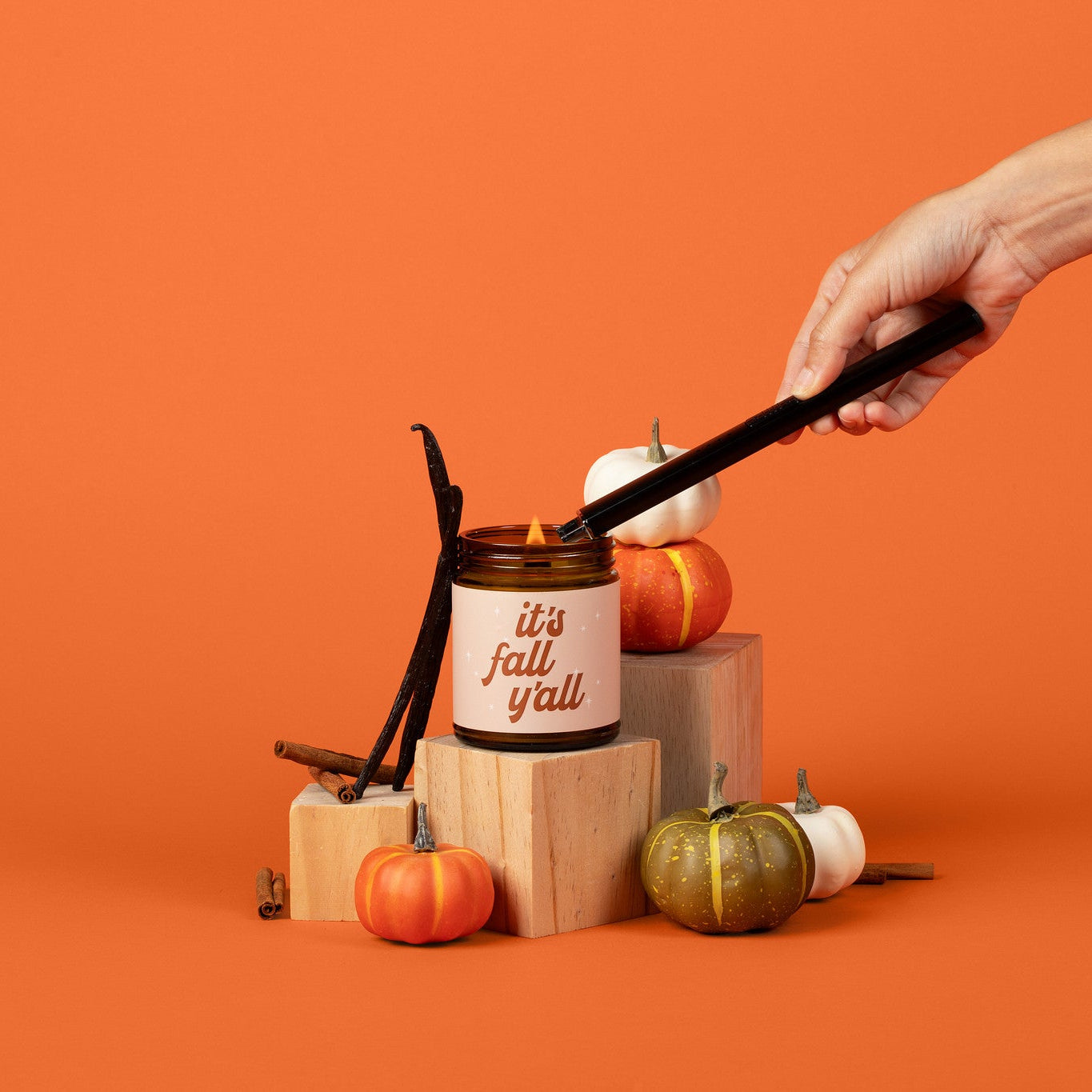 Hand using a lighter to light a jar candle labeled 'it's fall y'all' on an orange background with pumpkins.