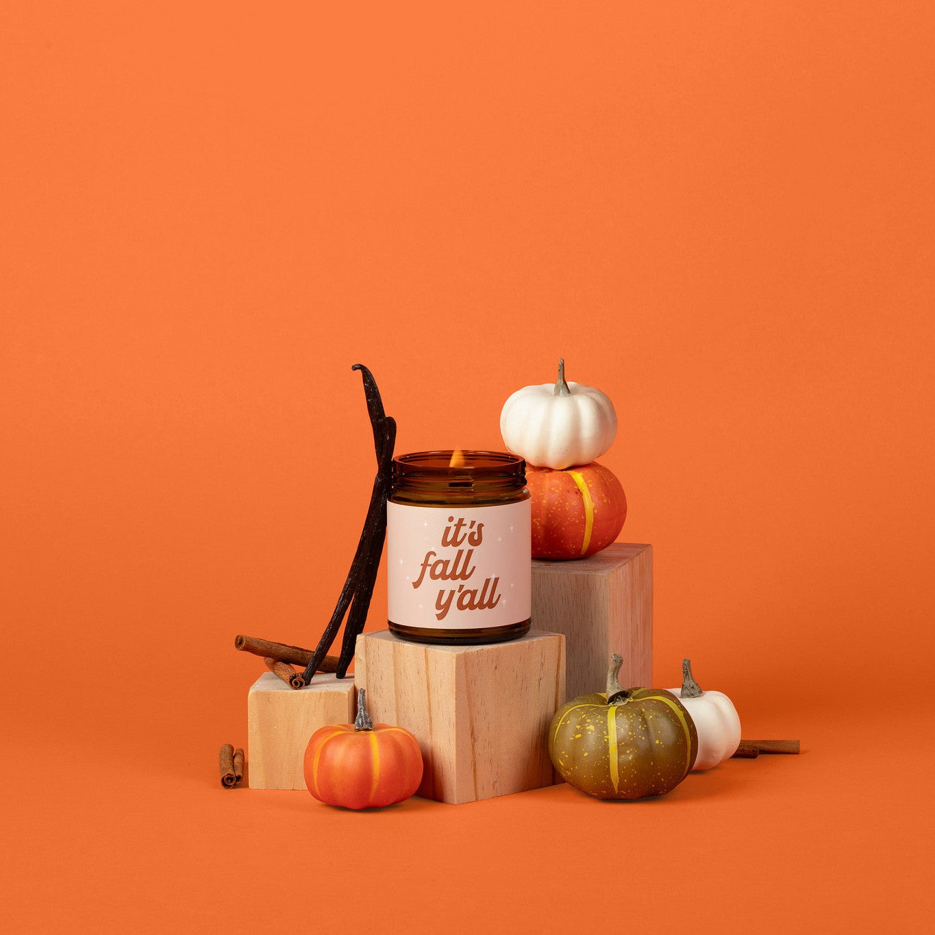 A fall-themed candle in an amber glass jar with a white label reading 'it's fall y'all', styled with small pumpkins, cinnamon sticks, and wood blocks against a solid orange background.