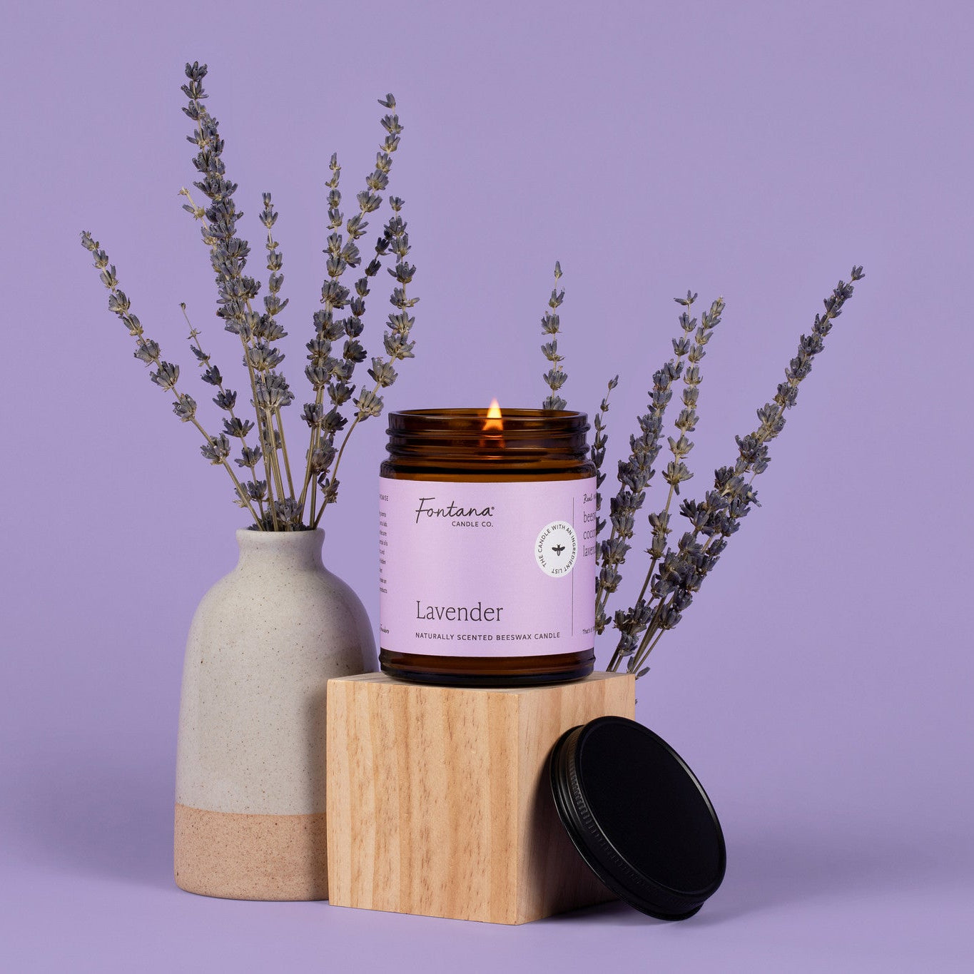 Lit amber glass Lavender jar candle with a light purple label, surrounded by Lavender bunches, in front of a darker purple background. 