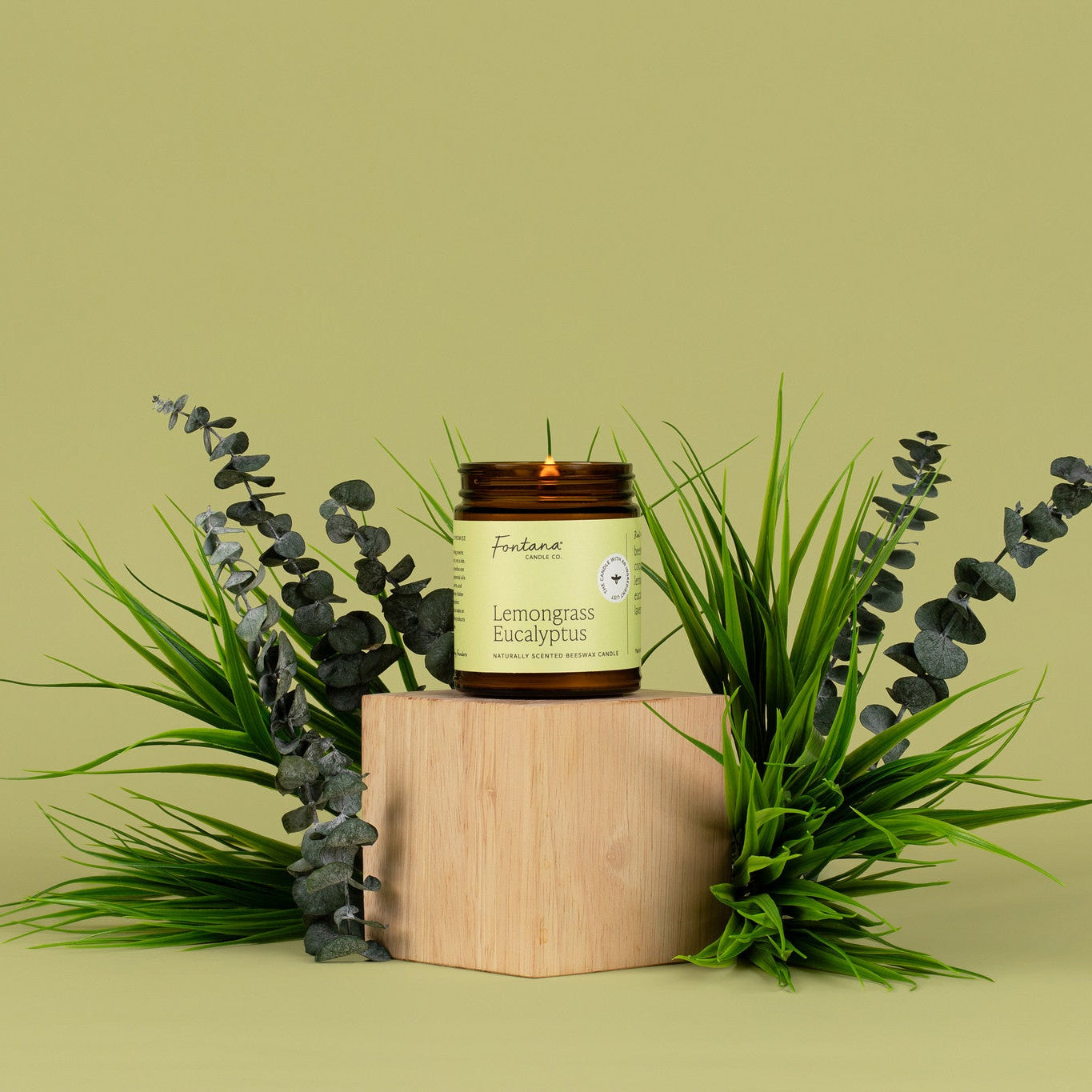 Lit amber glass candle labeled 'Lemongrass Eucalyptus' on a wooden block with greenery on a light green background.