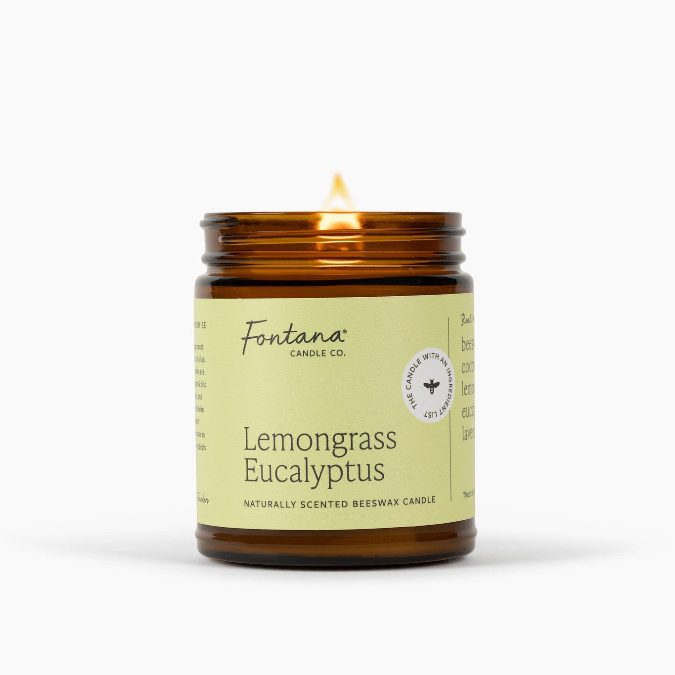 Lit amber glass jar candle with a light green/yellow label. The candle is in front of a white background. 