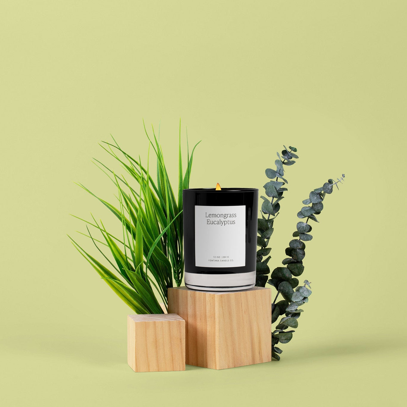 A black glass candle with a white label saying Lemongrass Eucalyptus sits on two light wood blocks, styled with green grass and eucalyptus stems against a solid pale yellow-green background.