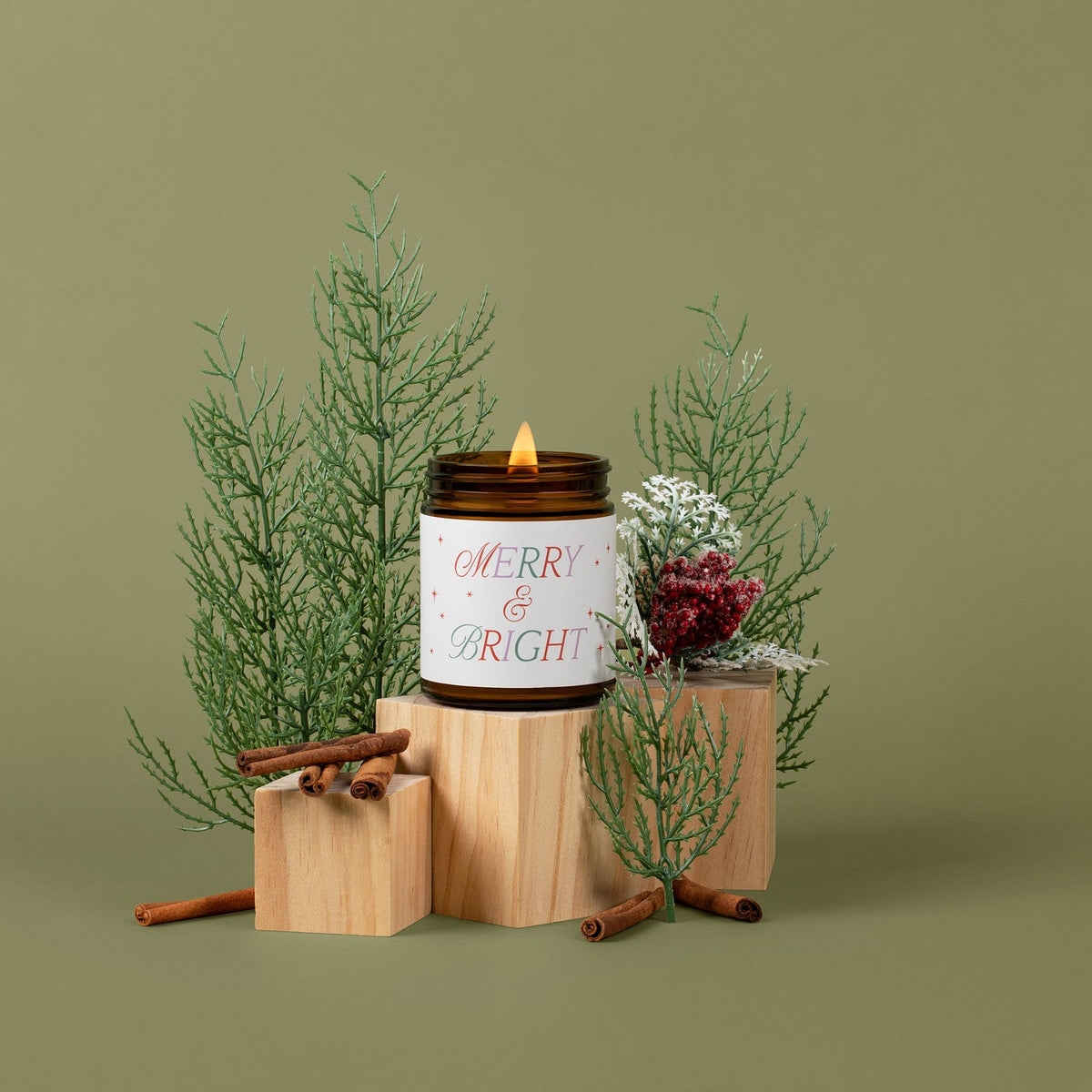 A lit candle in a dark amber glass jar with a white label reading 'MERRY &amp; BRIGHT' in red and green lettering, styled on wooden blocks with green pine branches, cinnamon sticks, and a small red berry sprig against a solid olive green background.