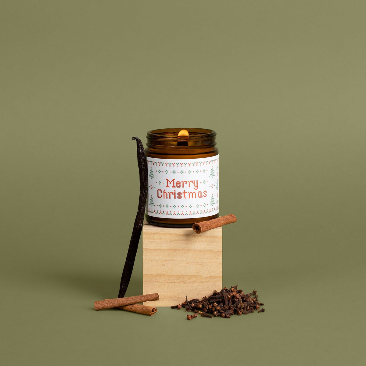 A holiday-themed candle in an amber glass jar with a white label reading 'Merry Christmas' in red text, styled on a wooden block with cinnamon sticks and dried cloves against an olive green background.