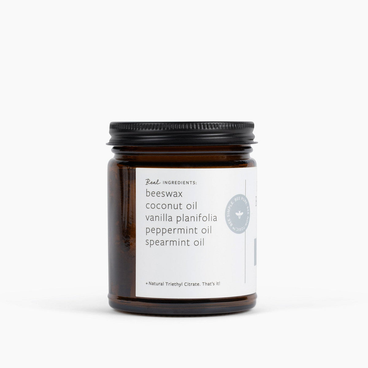 An amber glass jar candle with a black metal lid and a white label, photographed on a clean white background. The label reads 'Real Ingredients' and lists ingredients including beeswax, coconut oil, vanilla planifolia, peppermint oil and spearmint oil