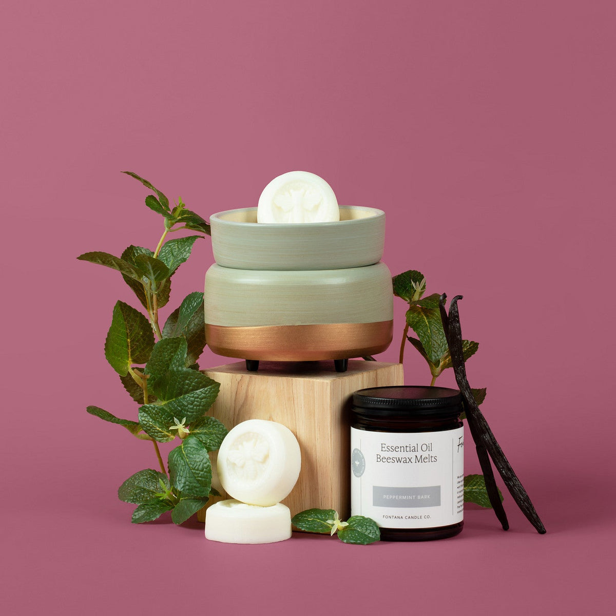 A styled product shot of a layered wax melt set on a wooden pedestal, featuring a sage green and copper-colored container with a white wax melt on top, alongside two white wax melts, green leaves, vanilla beans, and a small jar labeled 'Essential Oils &amp; Beeswax Melts Peppermint Bark' on a solid dusty pink background.