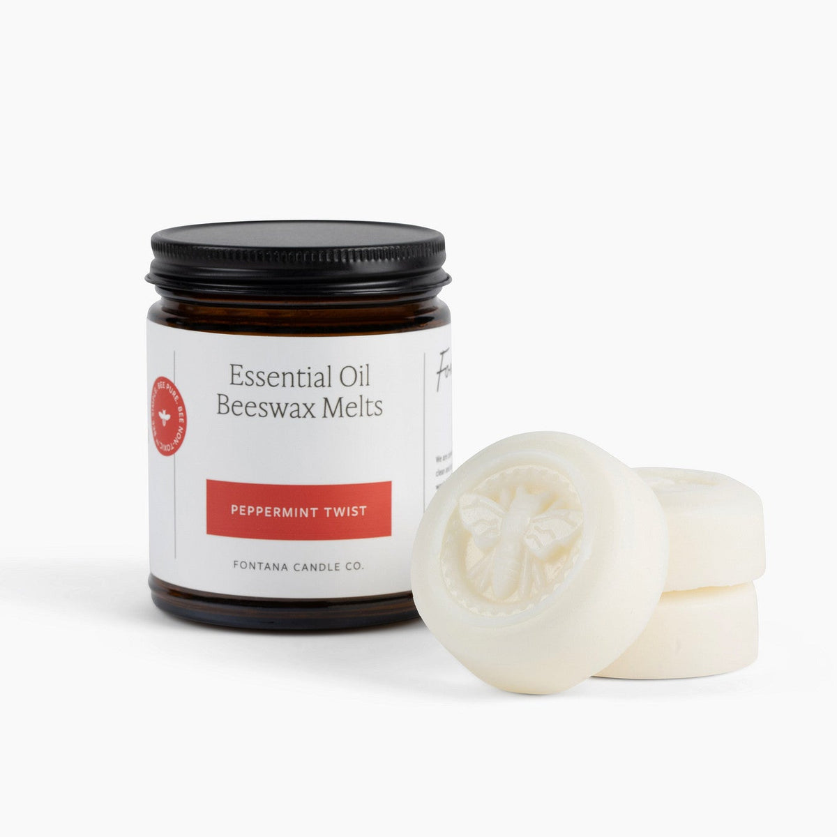 An amber glass jar labeled 'Essential Oil Beeswax Melts Peppermint Twist' with a black lid, shown next to three round off-white beeswax melts on a clean white background.