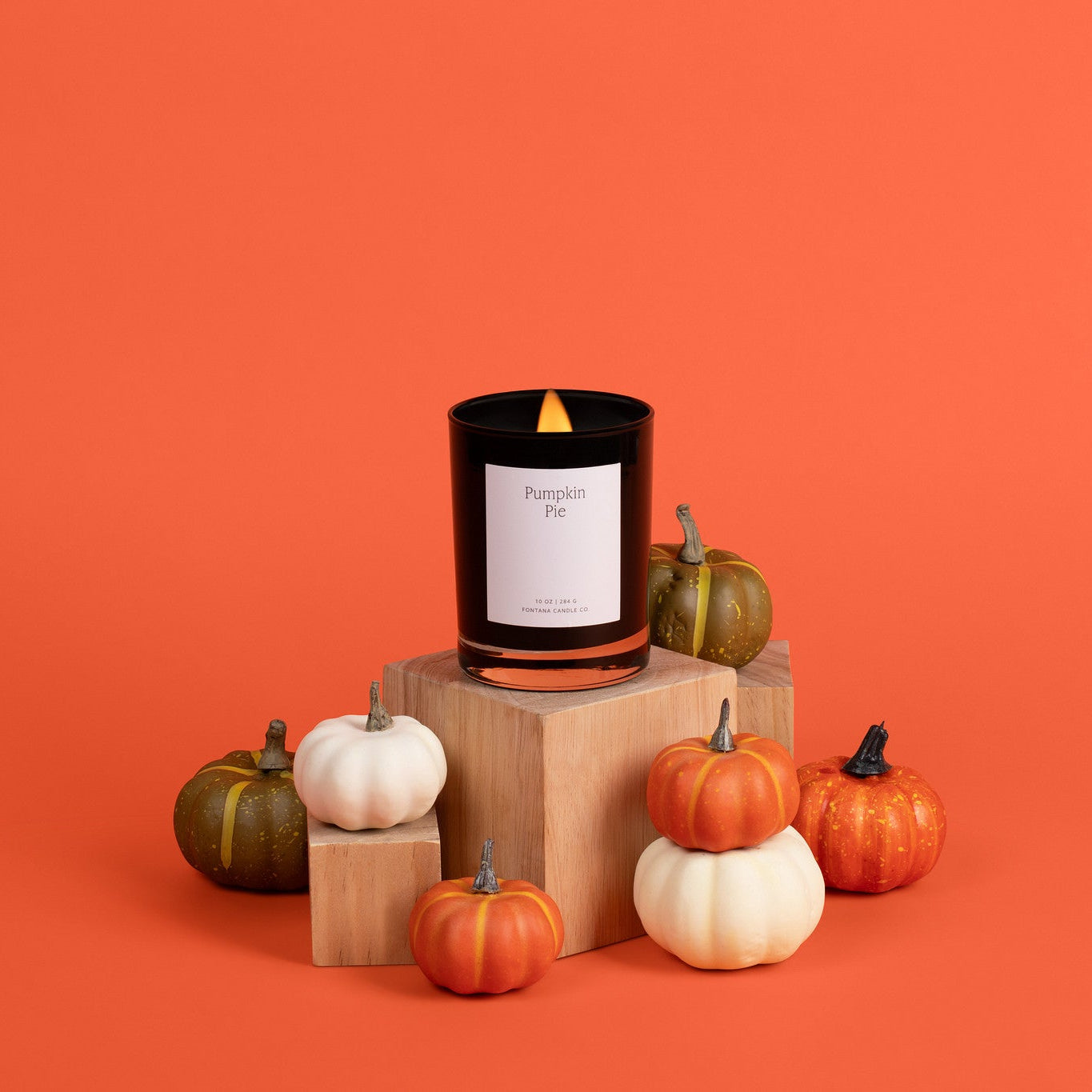 A black noir candle with a white label reading 'Pumpkin Pie' sits on a wooden block, surrounded by small orange, white, and green pumpkins against a solid orange background.