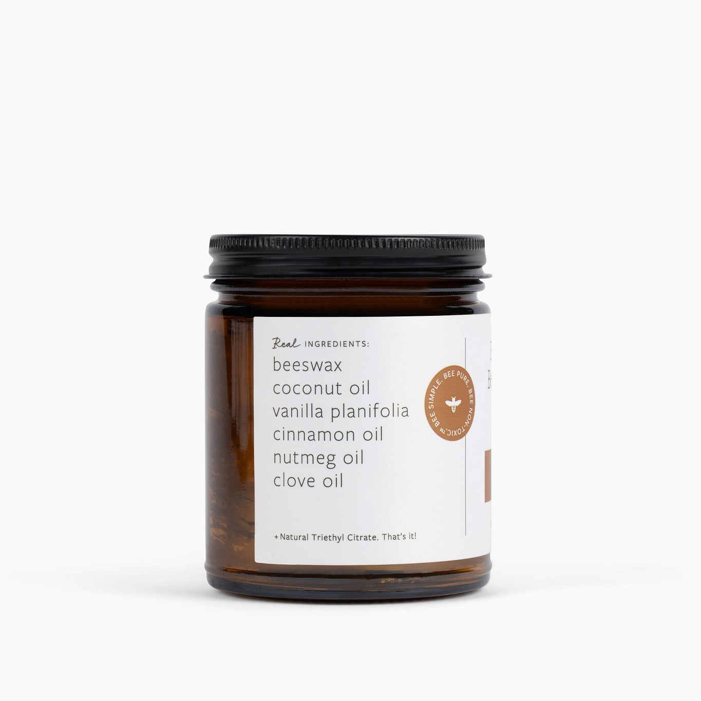 An amber glass jar candle with a black metal lid and a white label, photographed on a clean white background. The label reads 'Real Ingredients' and lists ingredients including beeswax, coconut oil, vanilla planifolia, cinnamon oil, nutmeg oil and clove oil