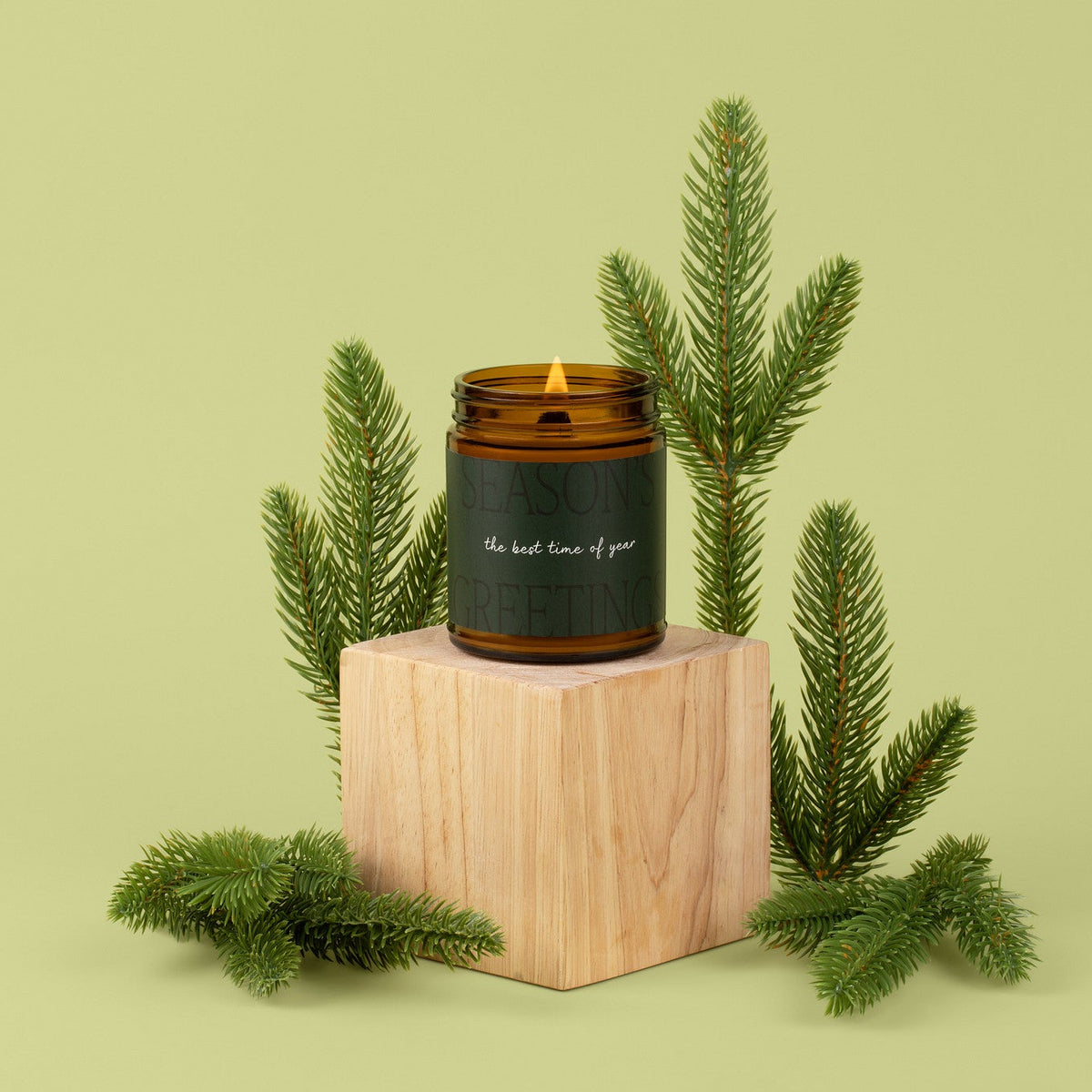 Glass amber jar candle with a dark green label sitting on top of wooden block surrounded by fir needle