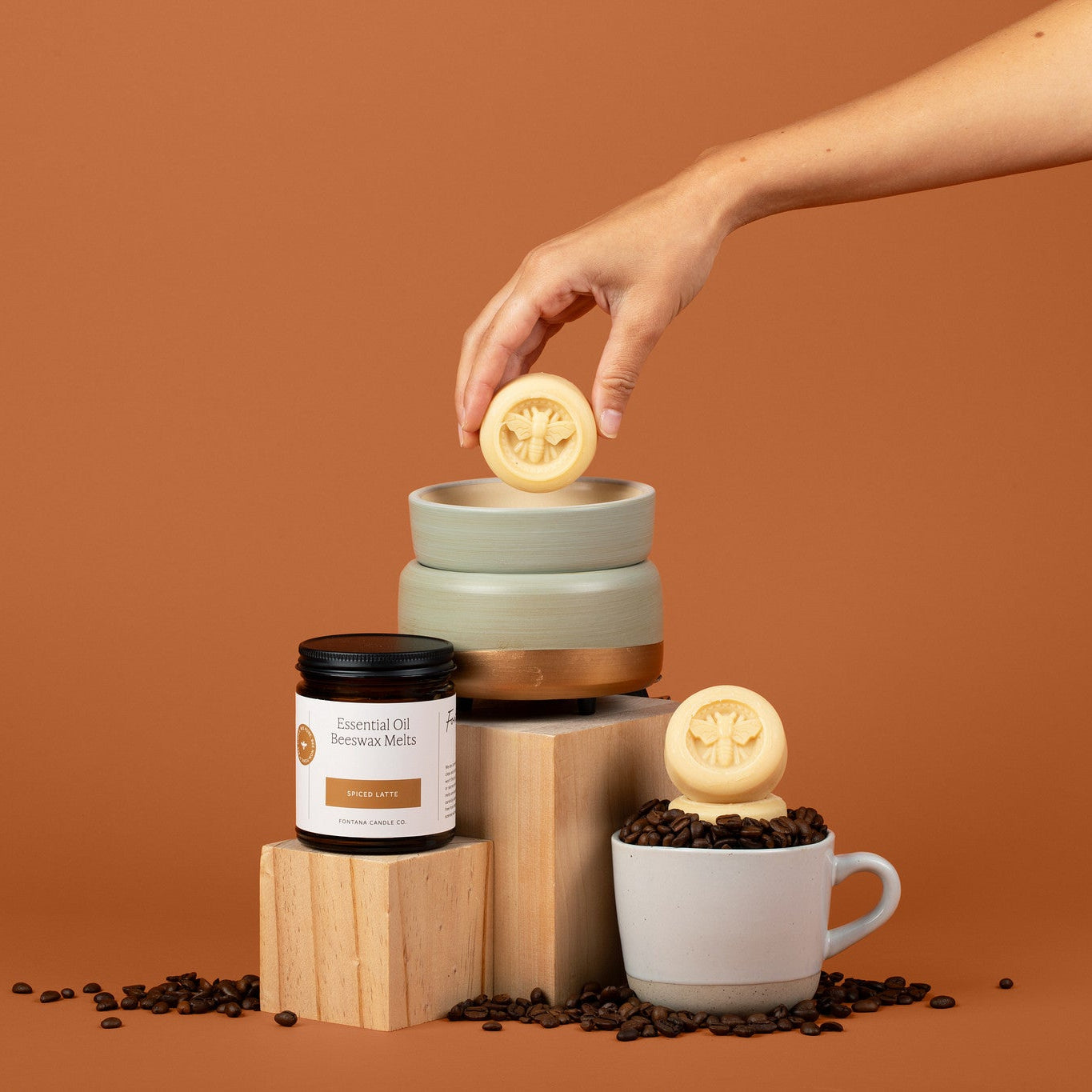 The teal wax melter on an orange background with the spiced latte wax melts in it. Surrounded by an assortment of coffee beans.