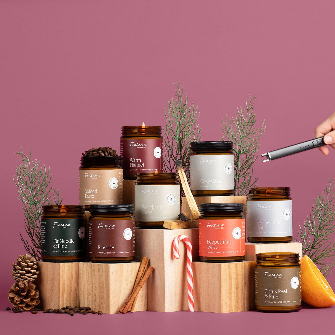 9 amber glass jar holiday candles in front of a burgendy background, surrounded by fir needles, candy canes, spices, and pinecones.