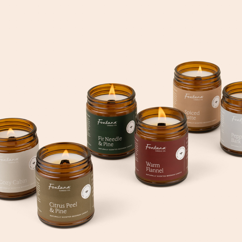Six amber glass jar candles with wooden wicks on a warm beige background. The labels show different scent names including Citrus Peel & Pine, Fir Needle & Pine, Warm Flannel, Spiced Latte, and Peppermint Bark.