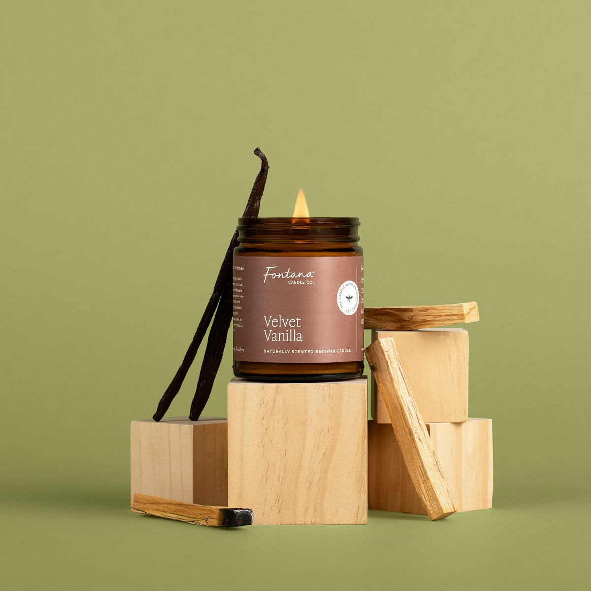 Velvet Vanilla scented amber glass jar candle with vanilla bean on wooden blocks against a green background