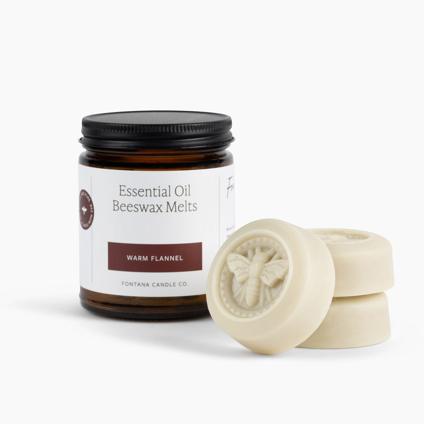 An amber glass jar labeled 'Essential Oil Beeswax Melts, Warm Flannel' with a black lid, shown next to three round off-white beeswax melts on a clean white background