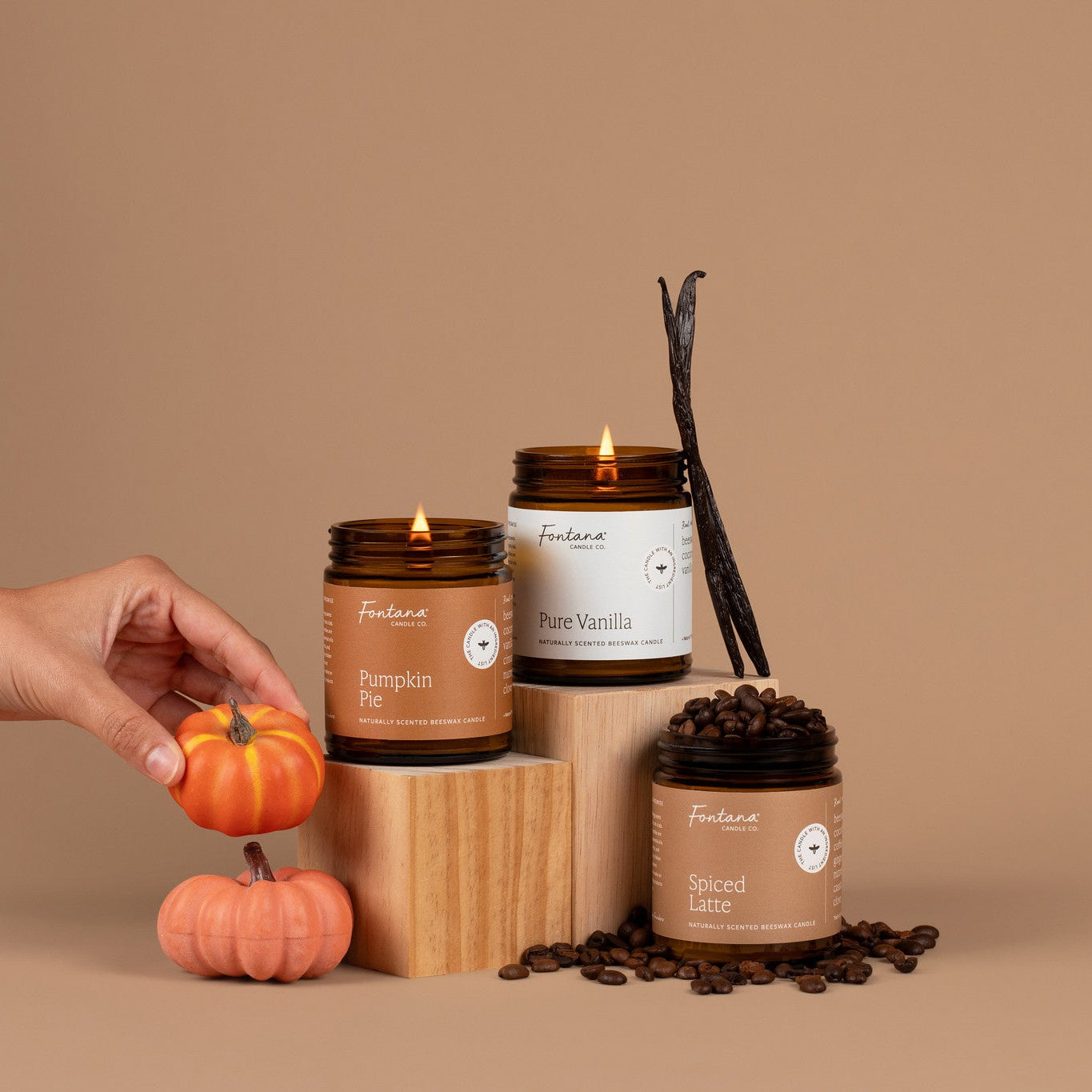 Candles with visible labels on a brown background, accompanied by pumpkins and coffee beans.